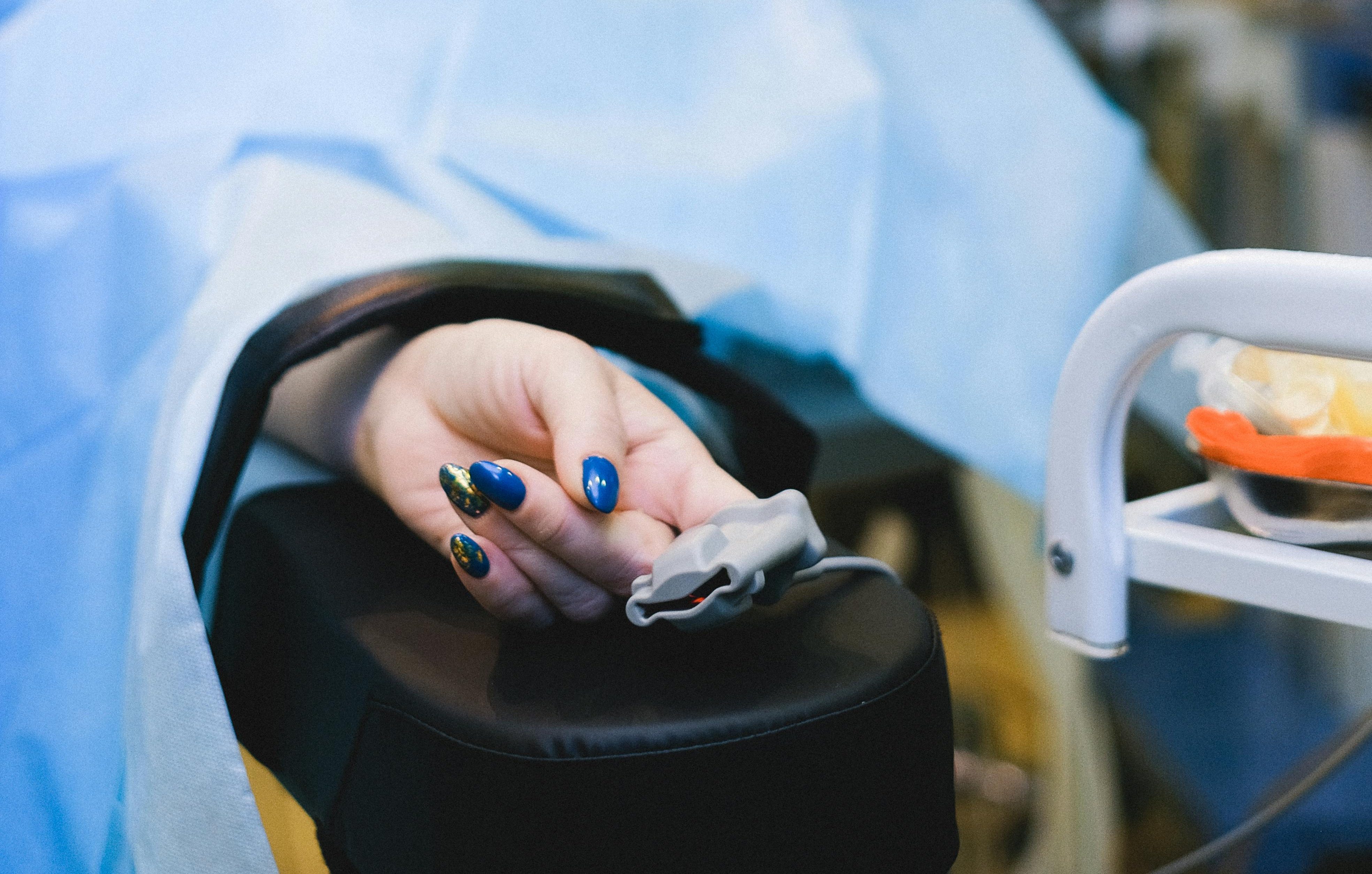 Alt text：在医疗环境中，病人的手放在扶手上，手指上夹着脉搏血氧仪。病人的指甲涂成蓝色和金色。背景是一面茶色的墙，墙上挂着医疗设备，包括一台显示生命体征的显示器。这与作品的主题 "科技如何改变急救医疗 "不谋而合。